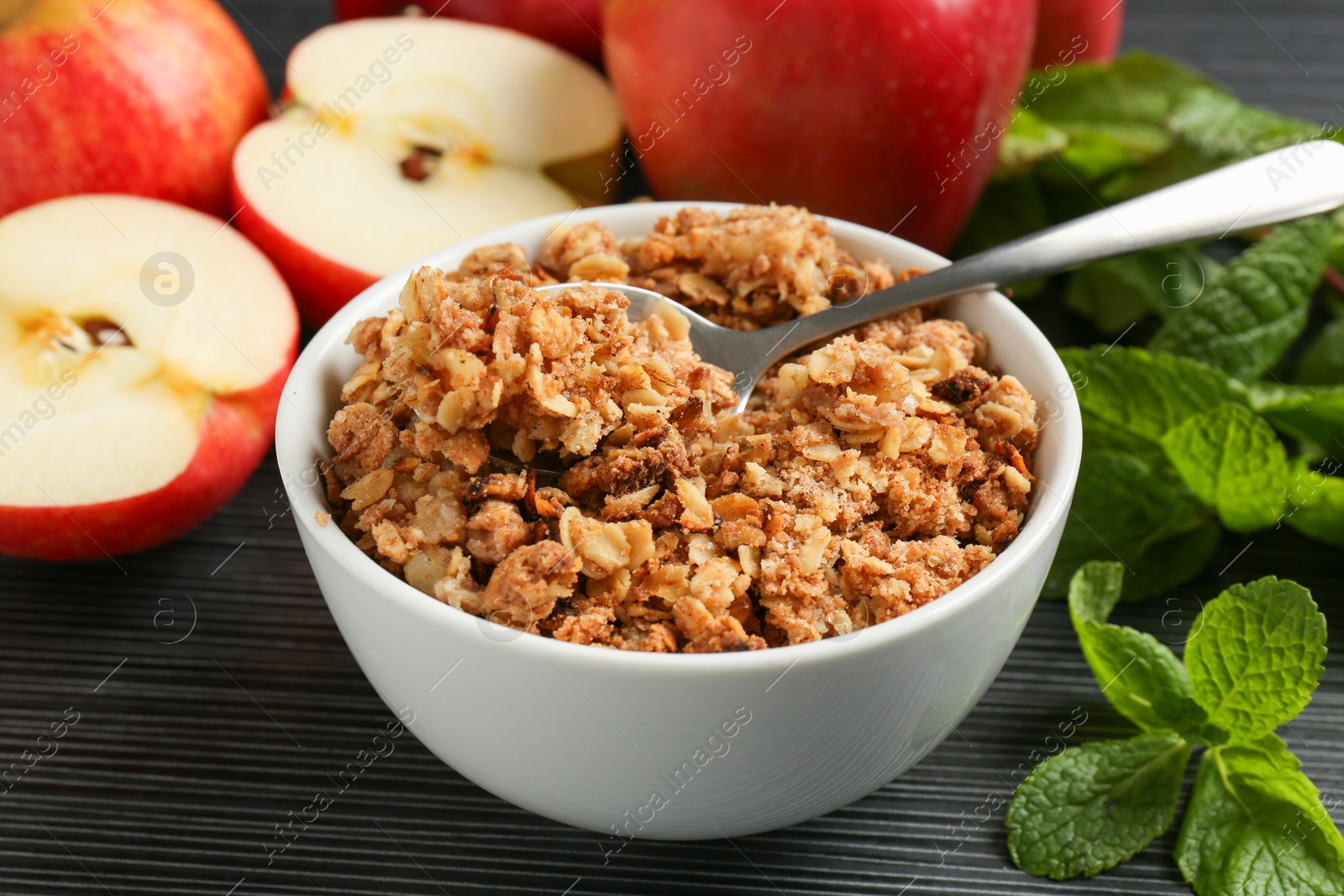 Photo of Delicious apple crisp in bowl, fresh fruits and mint on black textured table, closeup