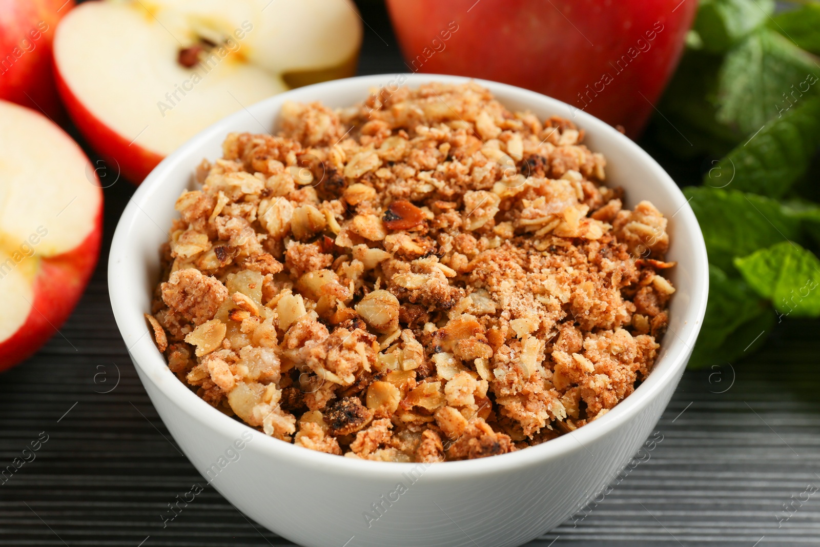 Photo of Delicious apple crisp in bowl, fresh fruits and mint on black textured table, closeup