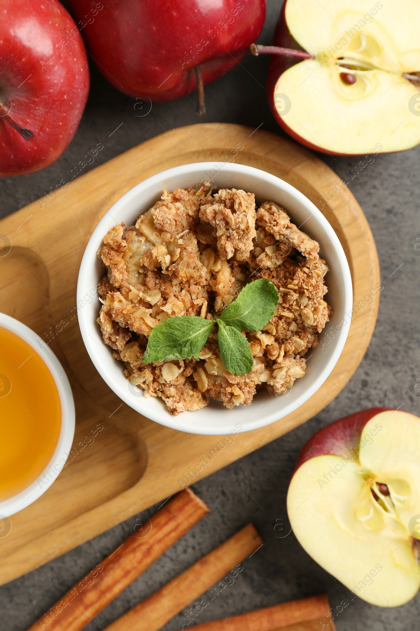 Photo of Delicious apple crisp in bowl, fresh fruits, cinnamon sticks, honey and mint on grey table, flat lay