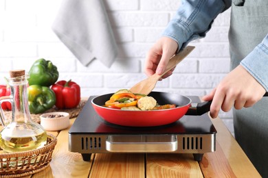 Photo of Woman frying vegetables in pan on stove at wooden table indoors, closeup