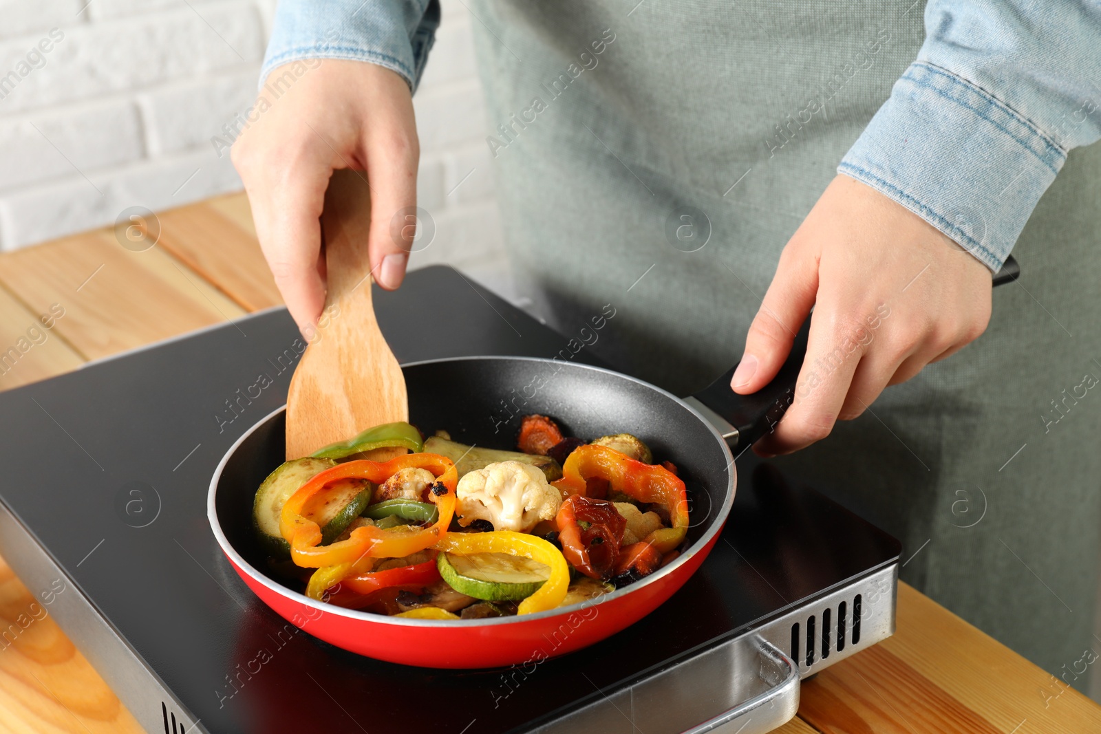 Photo of Woman frying vegetables in pan on stove at wooden table indoors, closeup