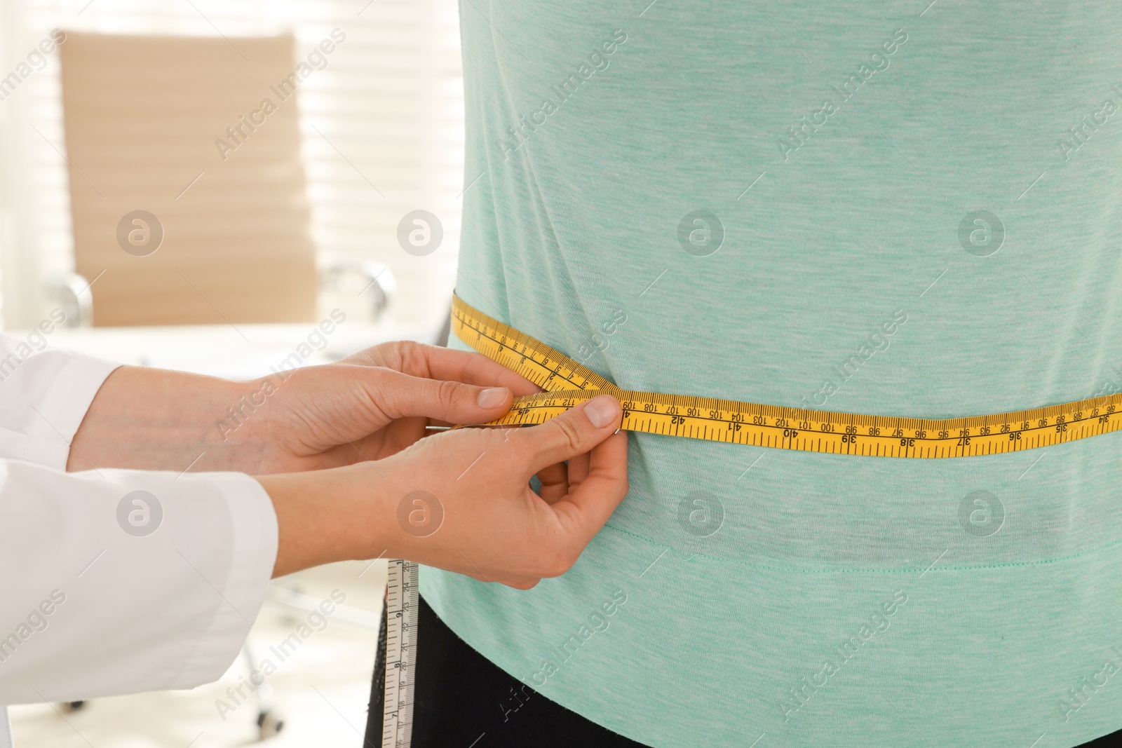 Photo of Nutritionist measuring overweight woman's waist with tape in hospital, closeup