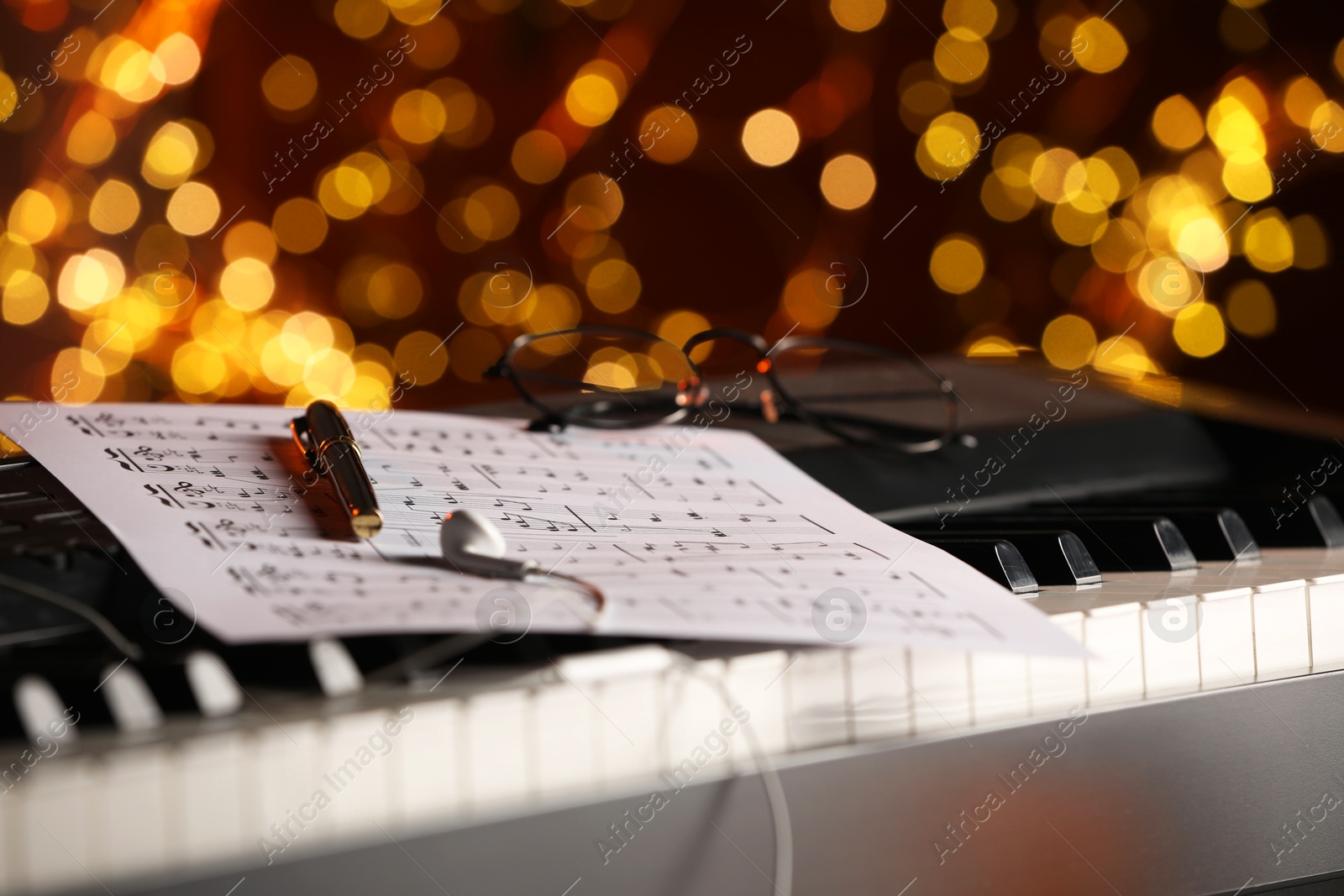 Photo of Sheet with musical notes, earphones and pen on piano against blurred lights, closeup. Bokeh effect