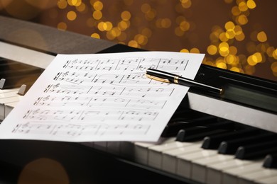 Photo of Sheet with musical notes and pen on piano against blurred lights, closeup. Bokeh effect