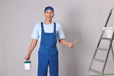 Photo of Smiling handyman with brush and bucket of paint near ladder indoors