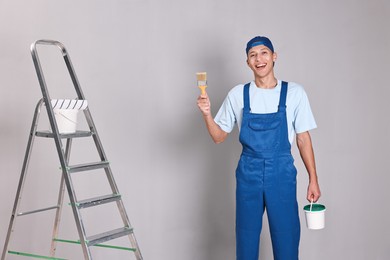 Photo of Smiling handyman with brush and bucket of paint near ladder indoors