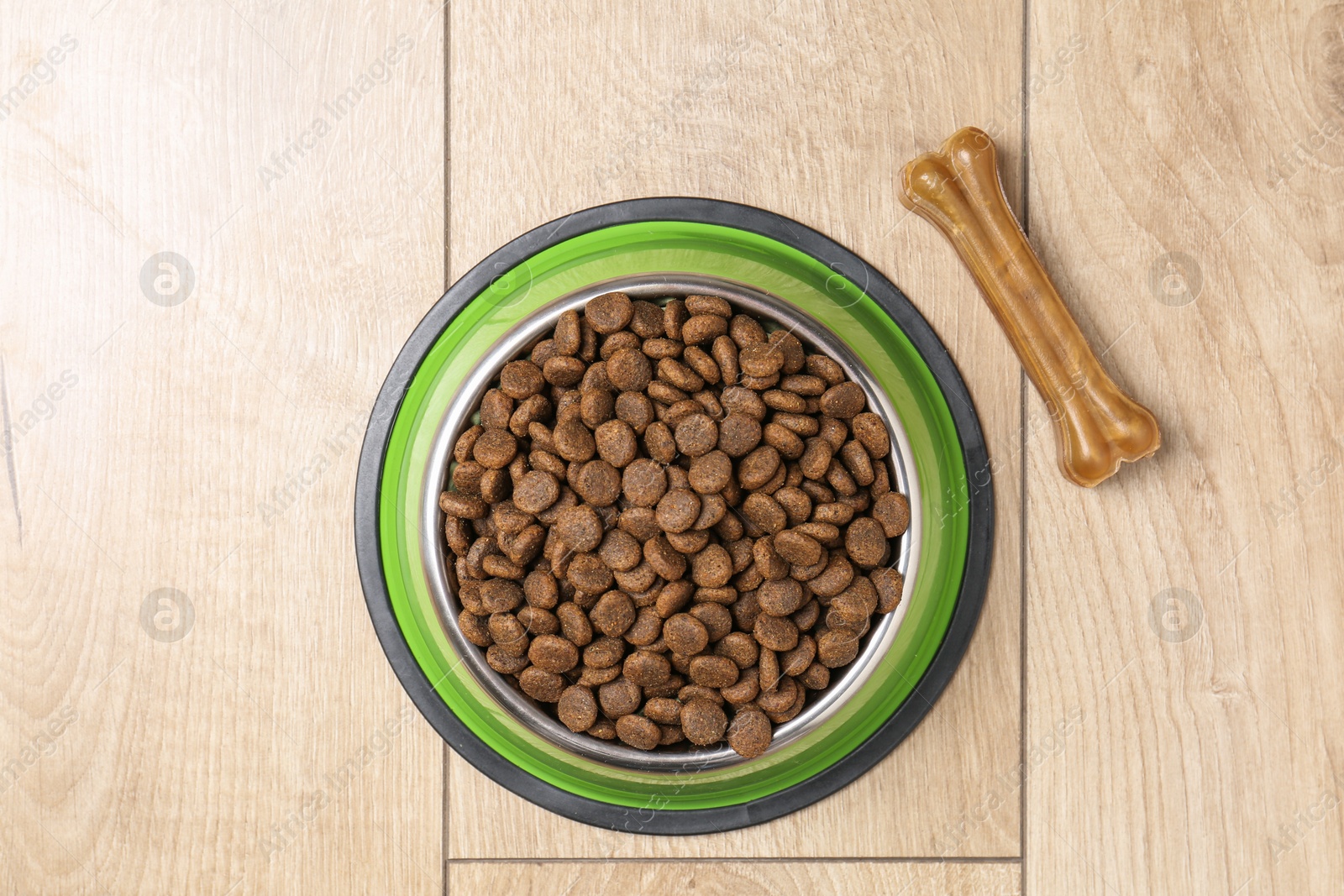 Photo of Dry pet food in feeding bowl and chew bone on wooden floor, top view