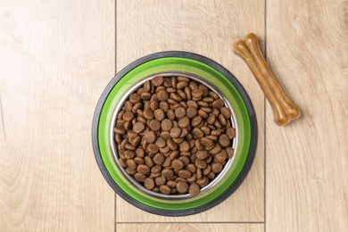 Photo of Dry pet food in feeding bowl and chew bone on wooden floor, top view