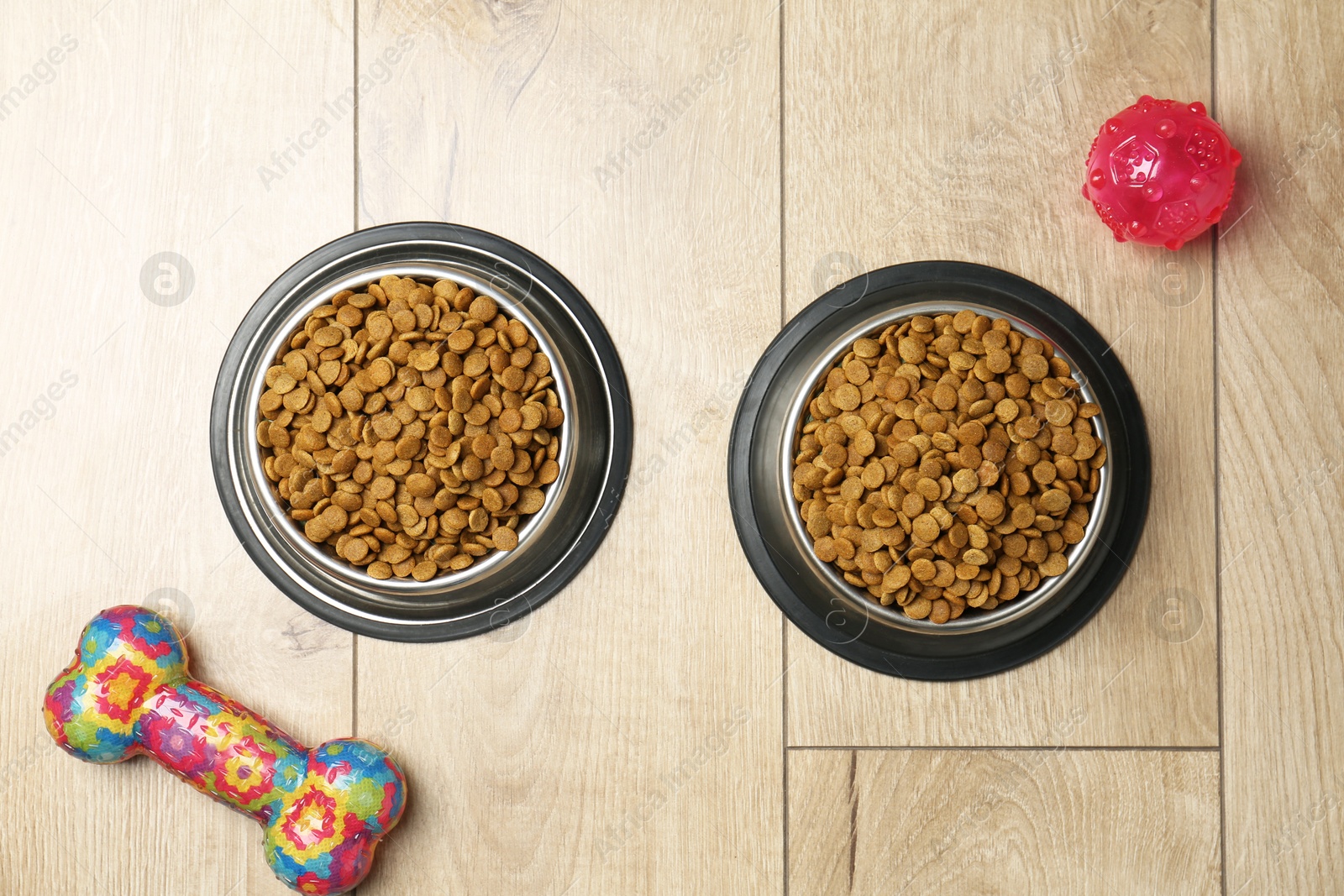 Photo of Dry pet food in feeding bowls and toys on wooden floor, flat lay