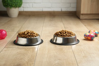 Photo of Dry pet food in feeding bowls and toys on floor indoors