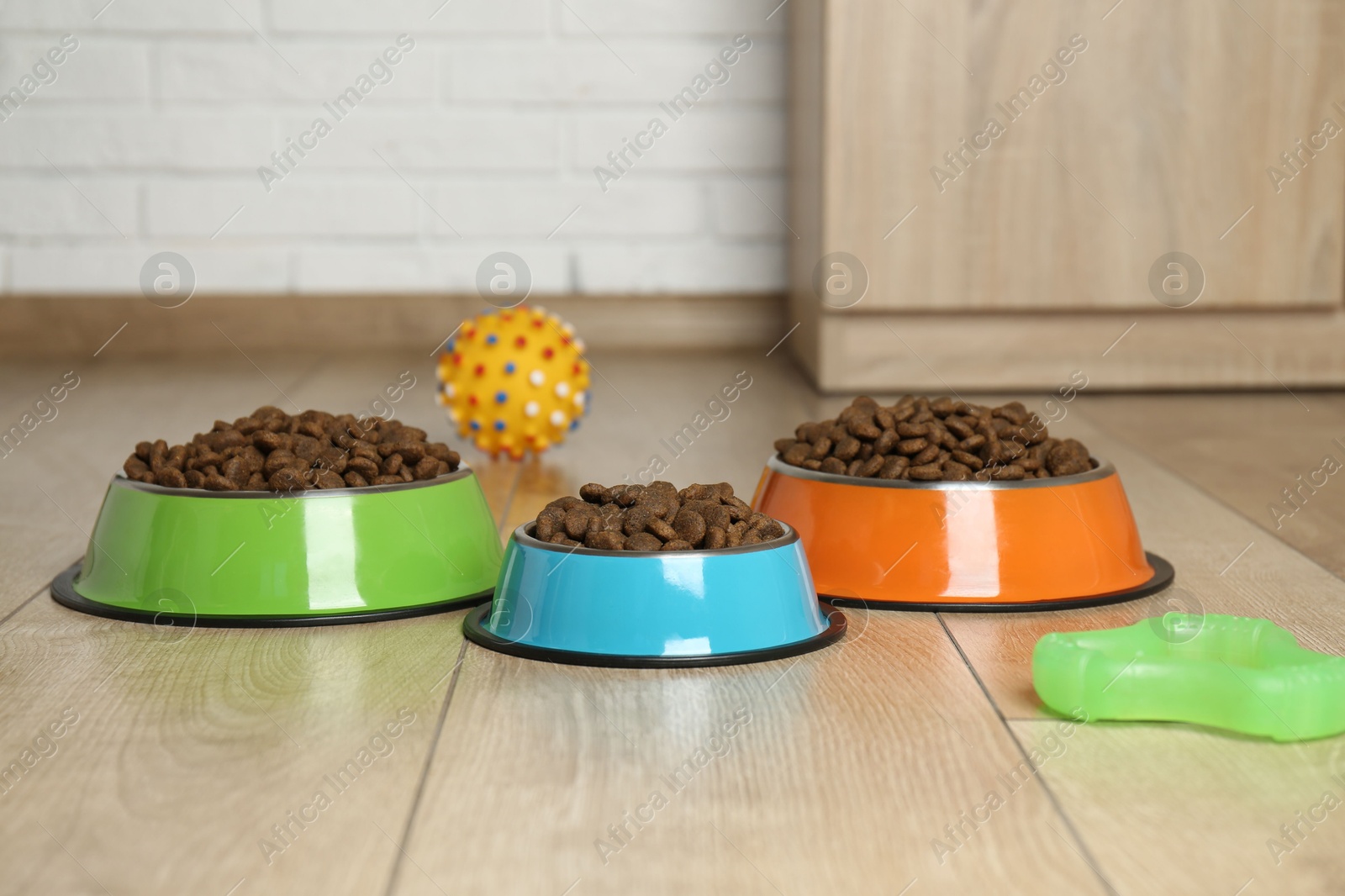 Photo of Dry pet food in feeding bowls and toys on floor indoors