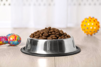 Photo of Dry pet food in feeding bowl and toys on floor indoors, closeup