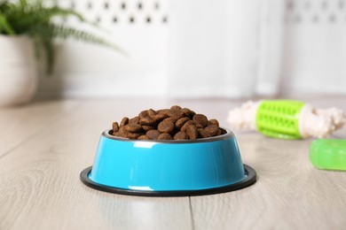 Photo of Dry pet food in feeding bowl and toys on floor indoors, closeup