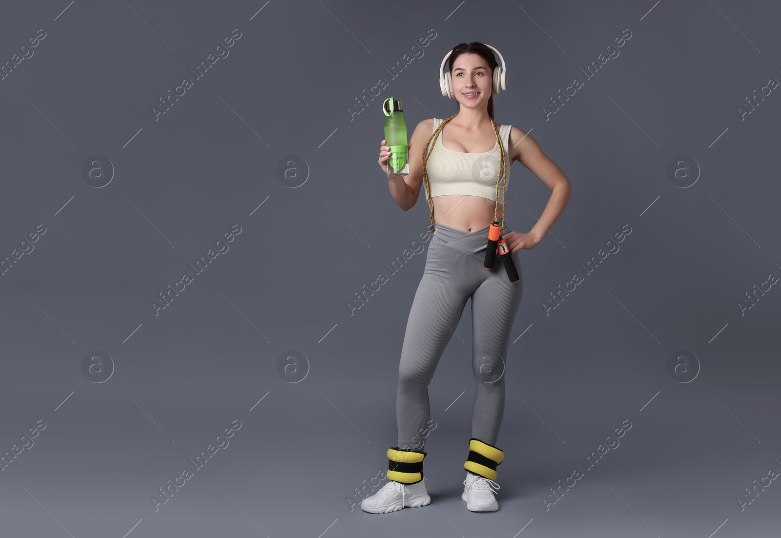 Photo of Woman in sportswear with headphones, bottle of water, ankle weights and skipping rope on grey background