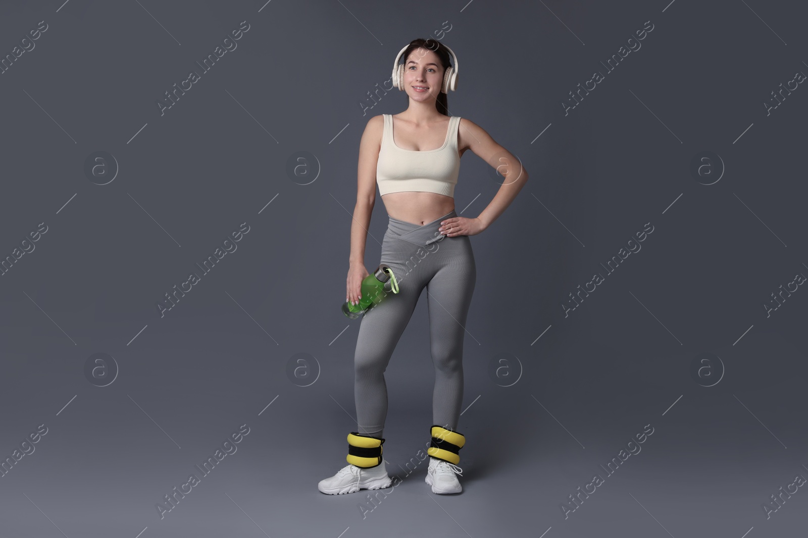 Photo of Woman in sportswear with headphones, bottle of water and ankle weights on grey background