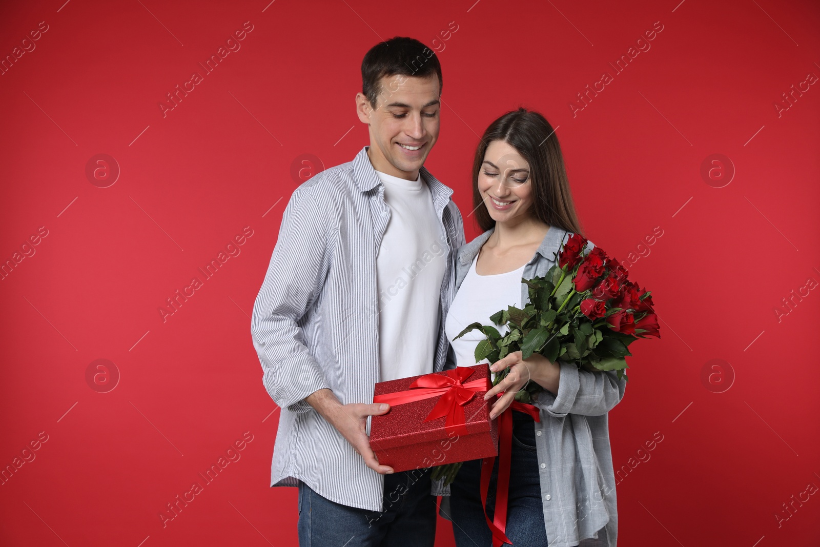 Photo of Lovely couple with bouquet of roses and gift box on red background. Valentine's day celebration