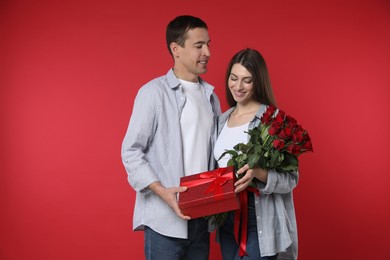 Photo of Lovely couple with bouquet of roses and gift box on red background. Valentine's day celebration