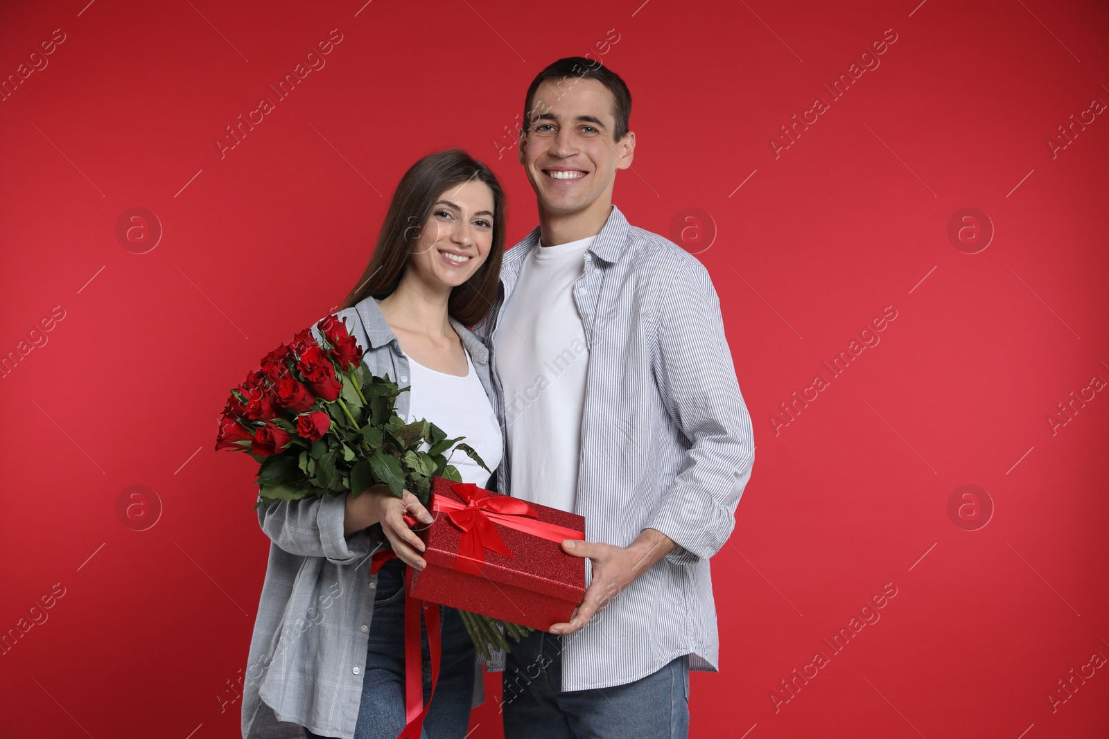 Photo of Lovely couple with bouquet of roses and gift box on red background. Valentine's day celebration