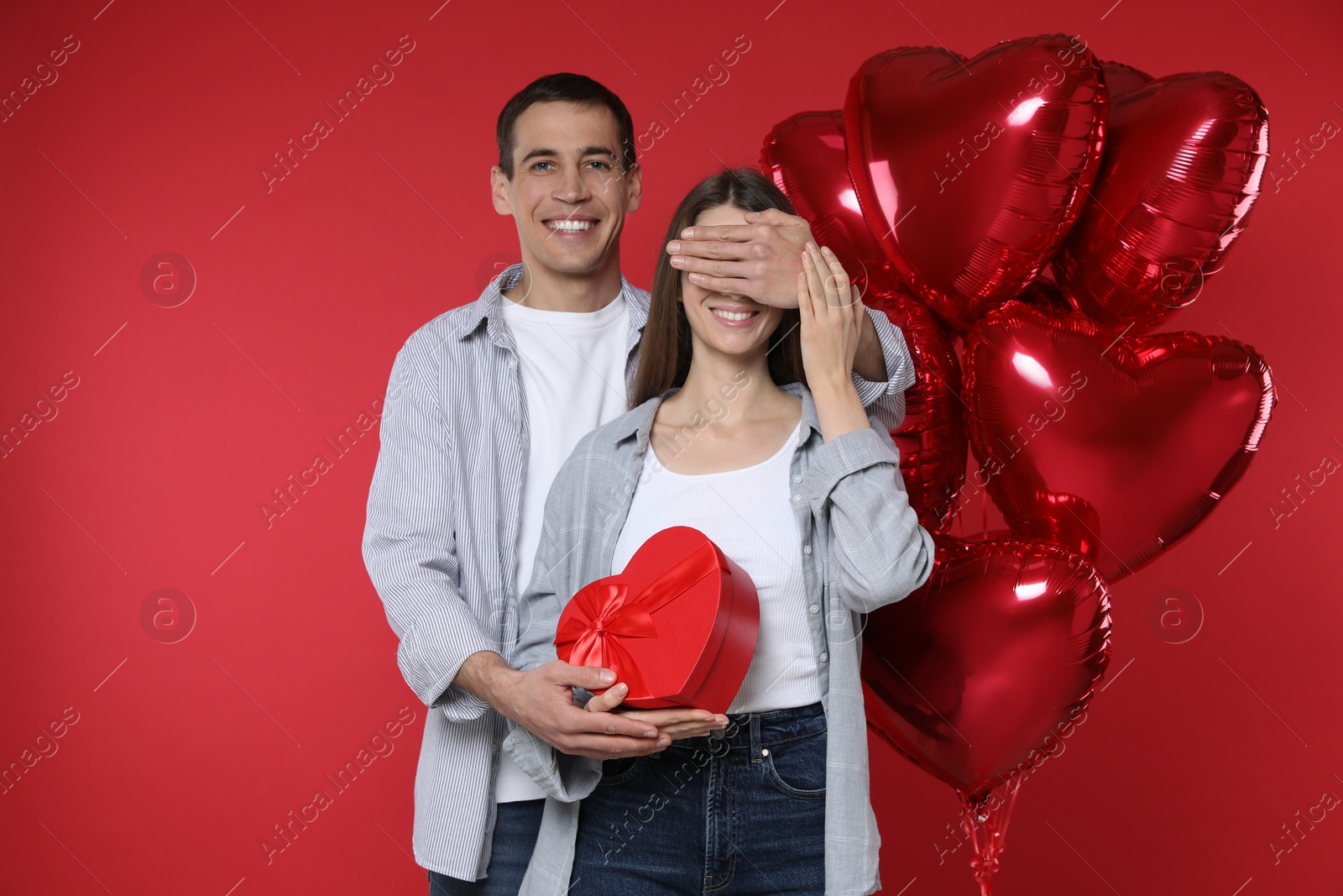 Photo of Lovely man surprising his girlfriend with Valentine's day gift near heart shaped balloons on red background