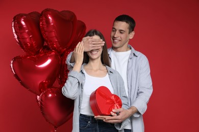 Photo of Lovely man surprising his girlfriend with Valentine's day gift near heart shaped balloons on red background