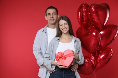 Photo of Lovely couple with gift box near heart shaped balloons on red background. Valentine's day celebration