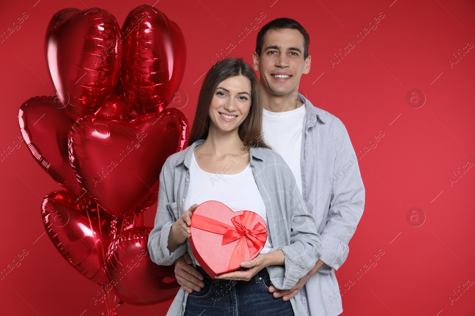 Photo of Lovely couple with gift box near heart shaped balloons on red background. Valentine's day celebration