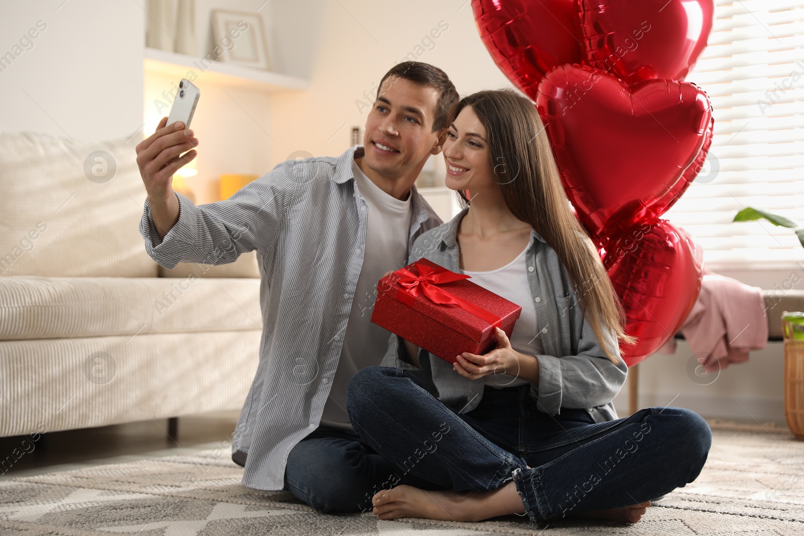 Photo of Lovely couple with gift box taking selfie near heart shaped balloons indoors. Valentine's day celebration