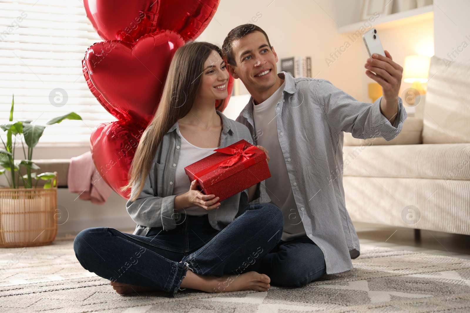 Photo of Lovely couple with gift box taking selfie near heart shaped balloons indoors. Valentine's day celebration