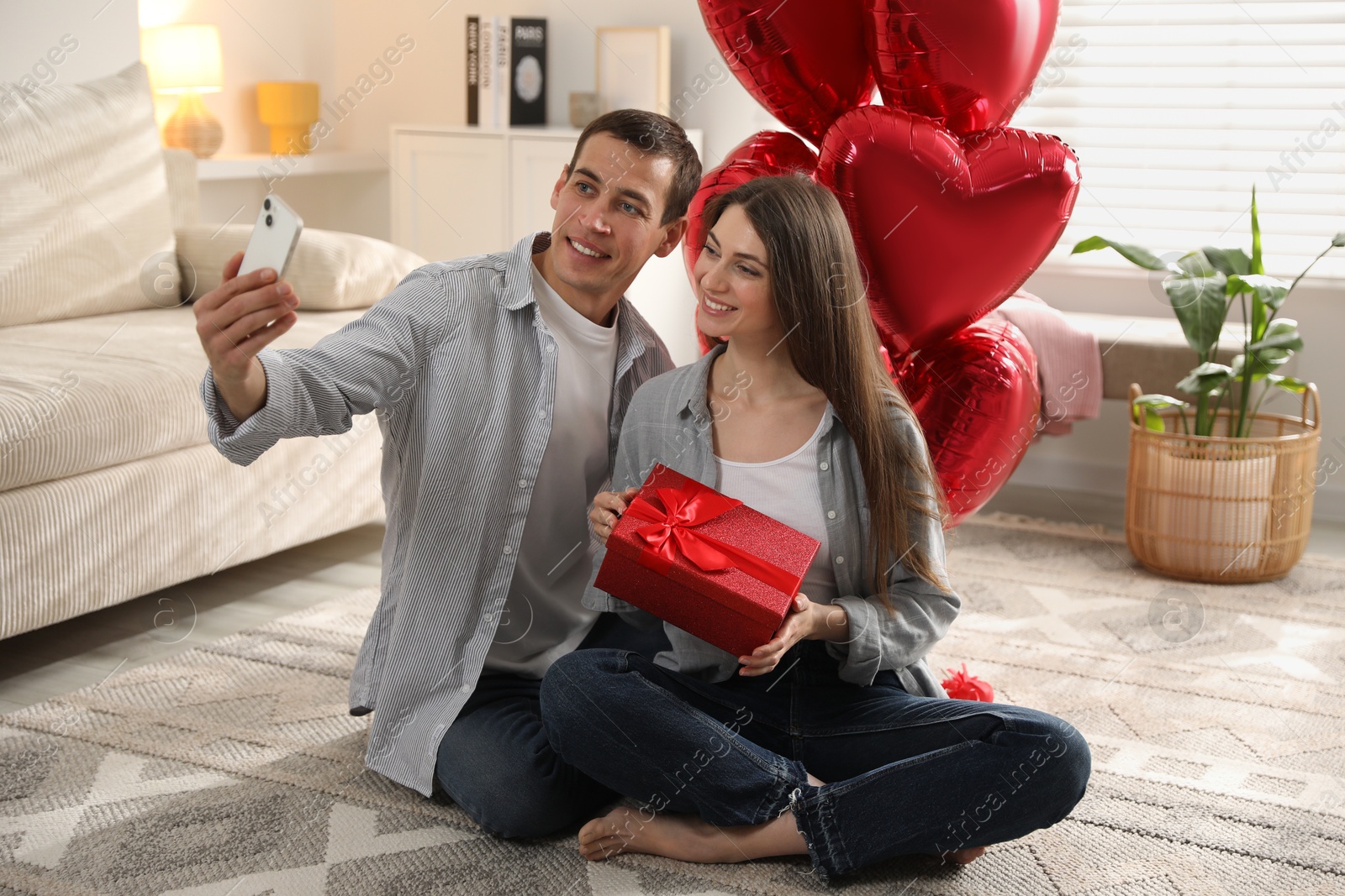 Photo of Lovely couple with gift box taking selfie near heart shaped balloons indoors. Valentine's day celebration