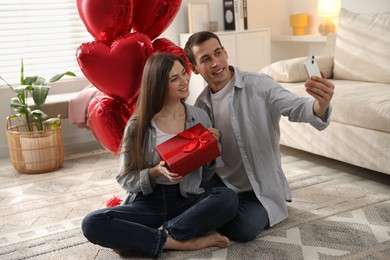 Photo of Lovely couple with gift box taking selfie near heart shaped balloons indoors. Valentine's day celebration