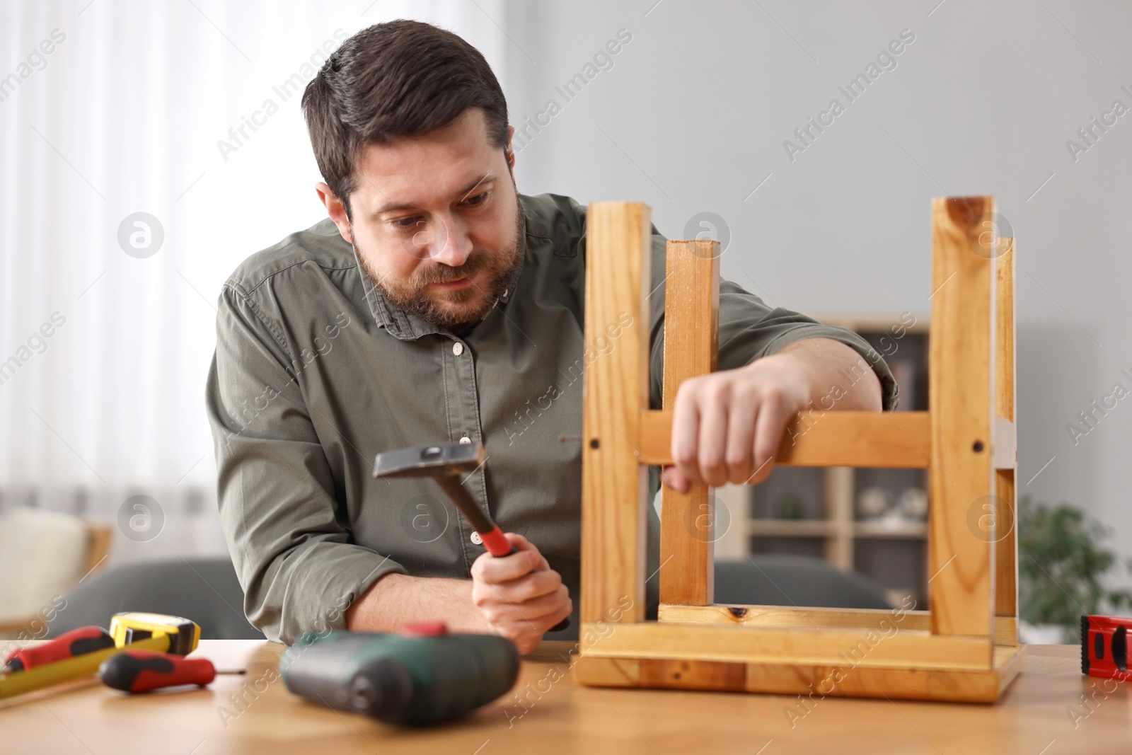 Photo of Man repairing wooden stool with nail and hammer at home