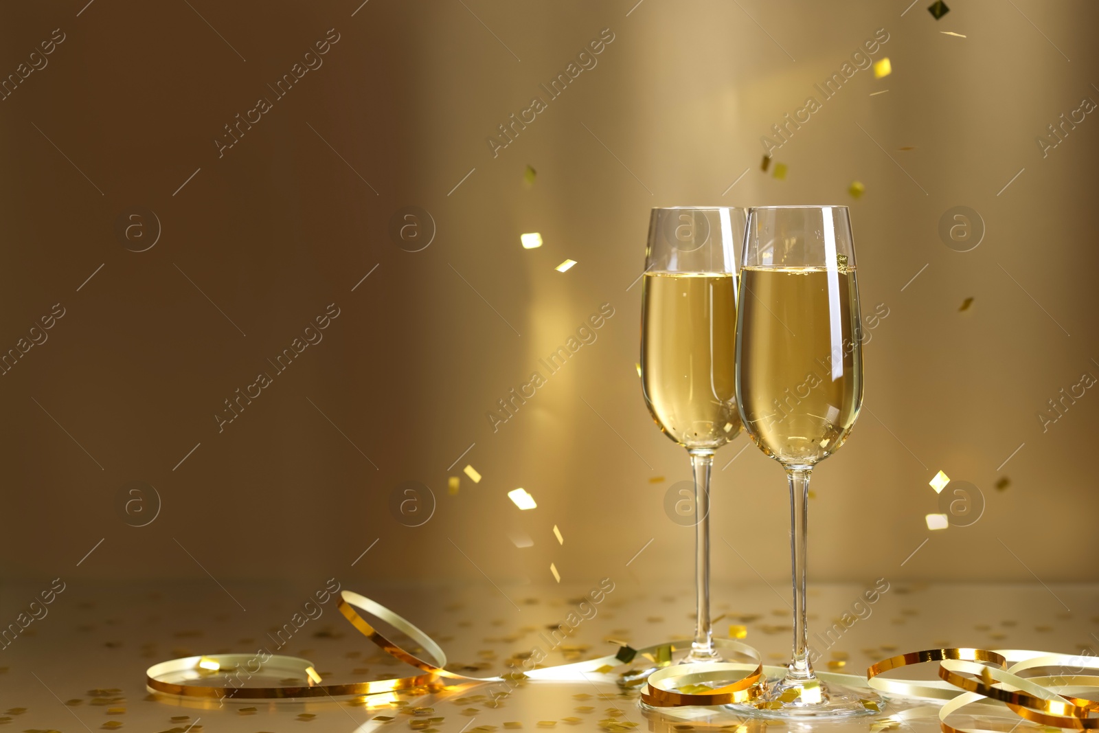 Photo of Champagne in glasses and flying confetti on golden background, space for text