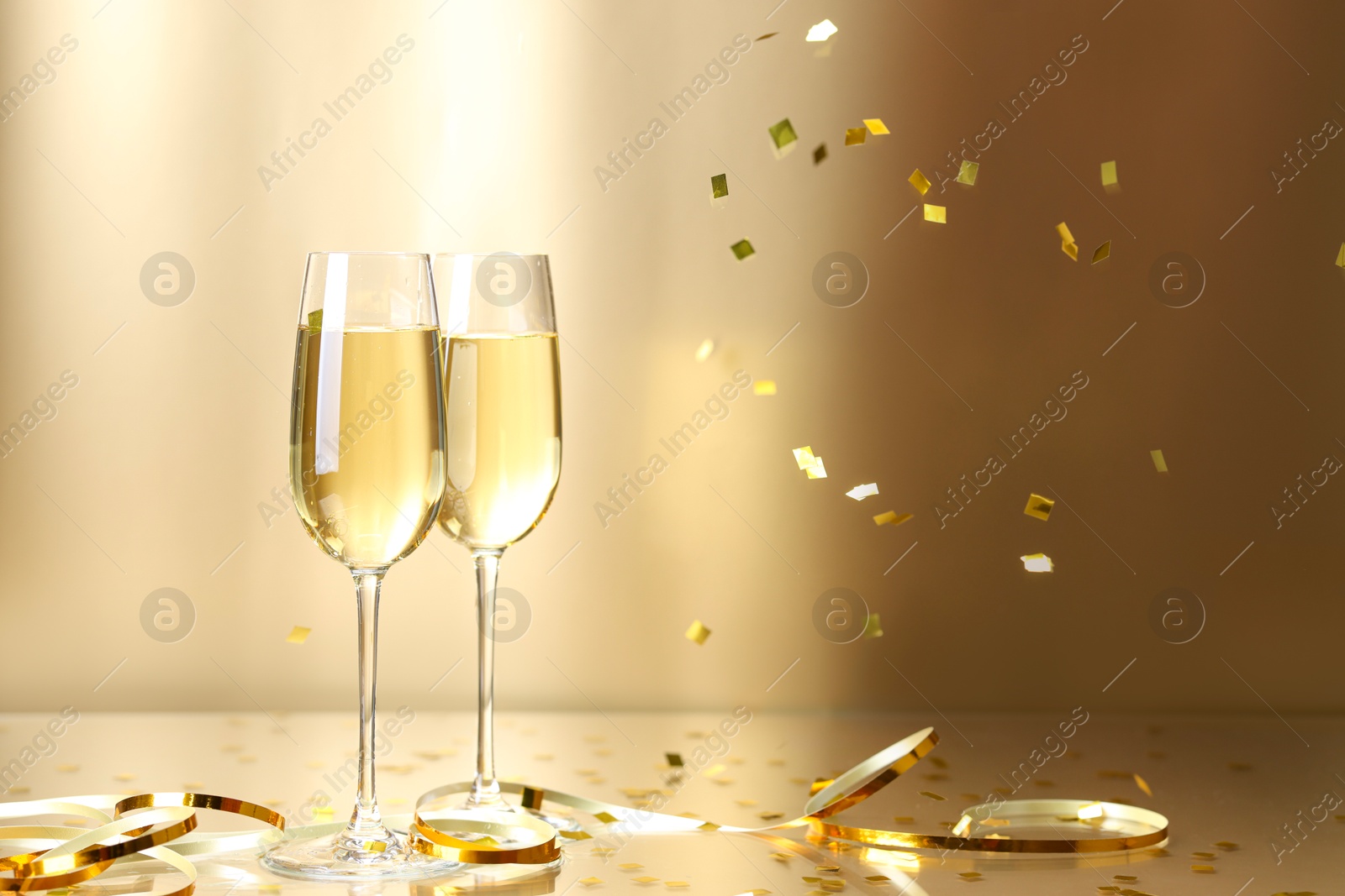 Photo of Champagne in glasses and flying confetti on golden background, space for text