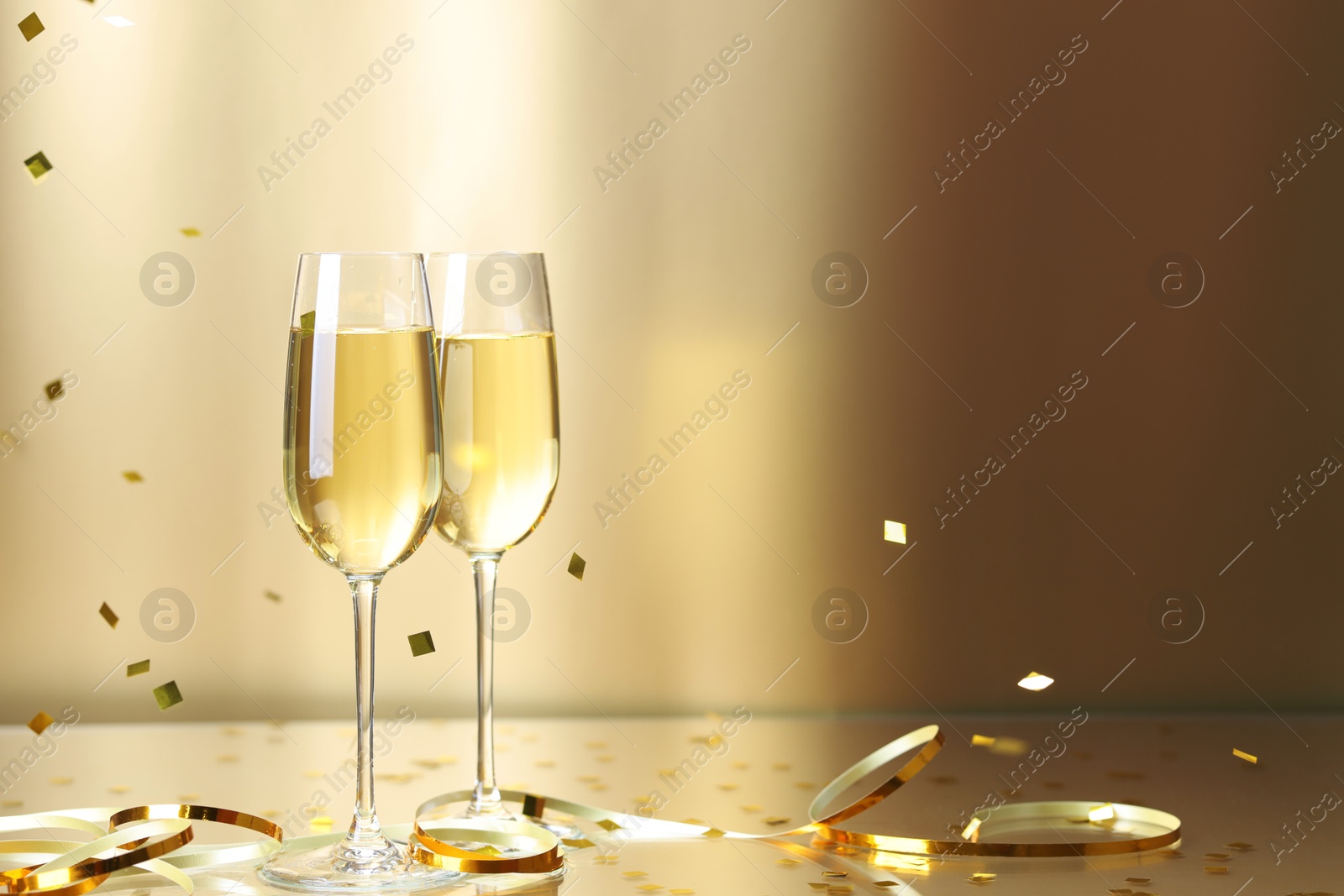 Photo of Champagne in glasses and flying confetti on golden background, space for text