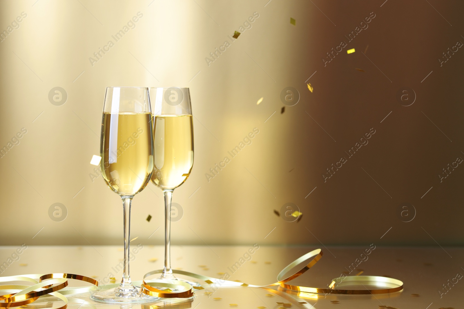 Photo of Champagne in glasses and flying confetti on golden background, space for text