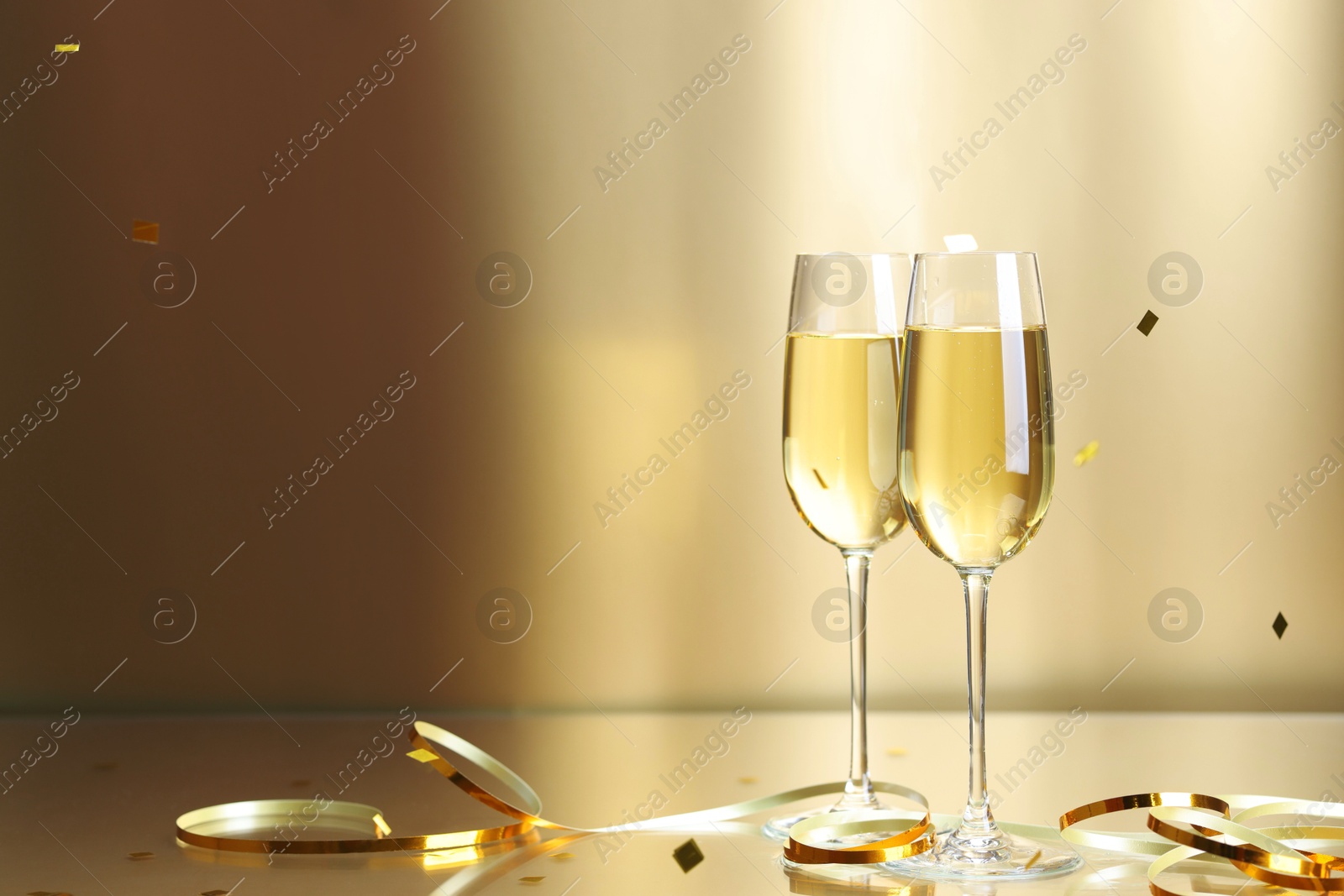 Photo of Champagne in glasses and flying confetti on golden background, space for text