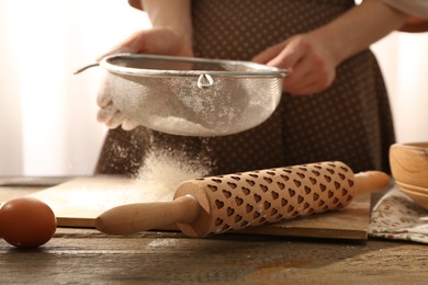 Photo of Woman sifting flour at wooden table with rolling pin indoors, closeup