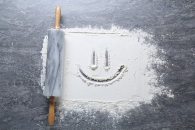 Photo of Rolling pin and smiling face made of flour on grey textured table, top view