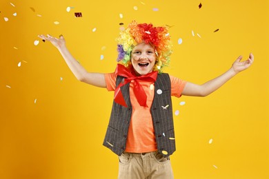 Photo of Happy little boy dressed like clown under falling confetti on orange background. Surprise party