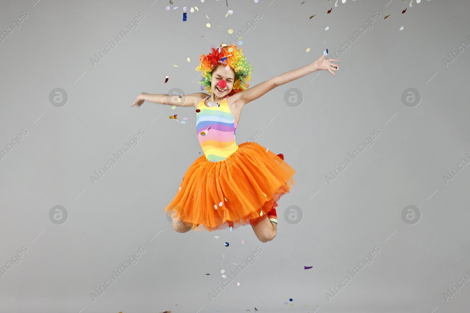 Photo of Girl dressed like clown jumping under flying confetti on grey background. Surprise party