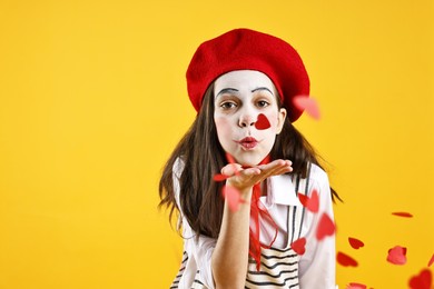 Photo of Cute girl dressed like mime blowing out confetti in shape of hearts on yellow background, space for text. Surprise party