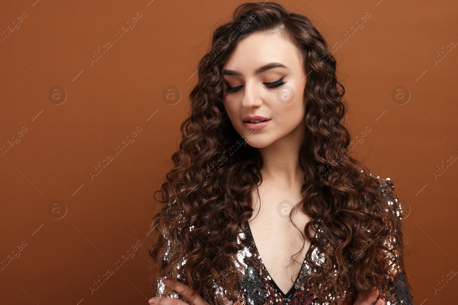 Photo of Beautiful young woman with long curly hair in sequin dress on brown background, space for text