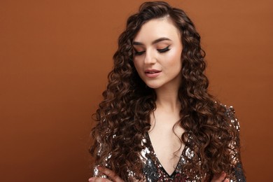 Photo of Beautiful young woman with long curly hair in sequin dress on brown background, space for text