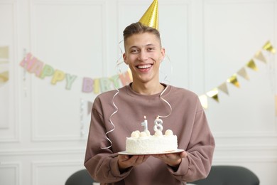 Photo of Coming of age party - 18th birthday. Happy young man in hat holding tasty cake with number shaped candles at home