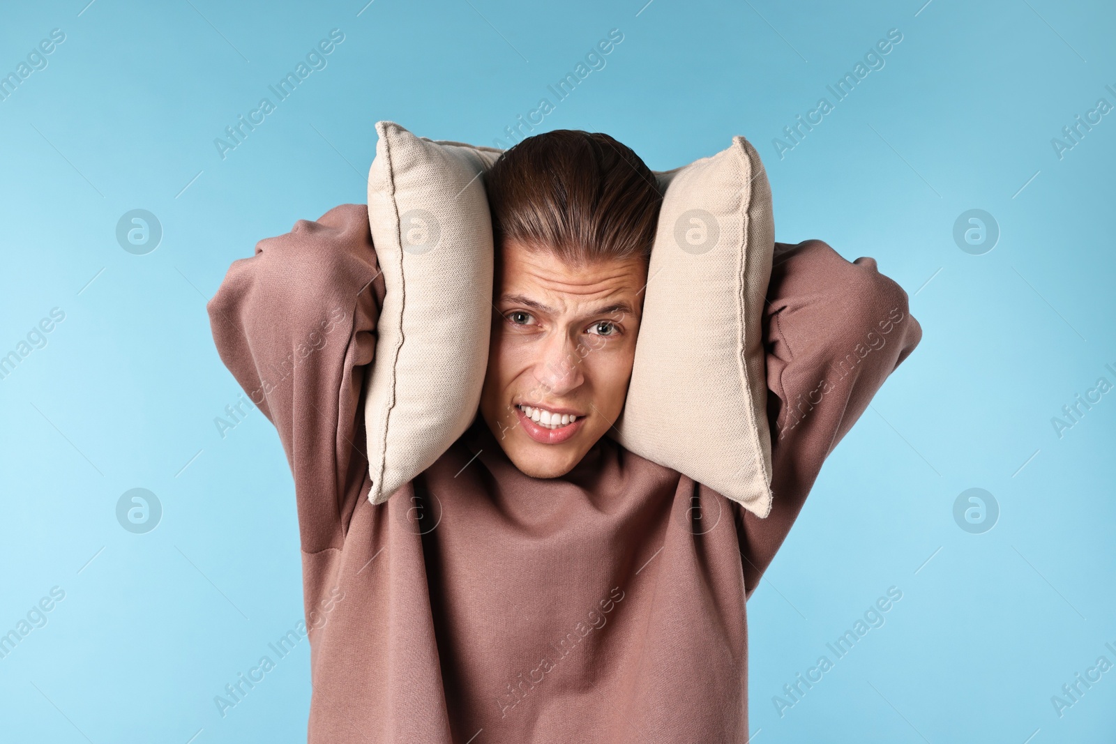 Photo of Annoyed young man covering his ears with pillows due to loud sound on light blue background