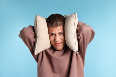 Photo of Annoyed young man covering his ears with pillows due to loud sound on light blue background