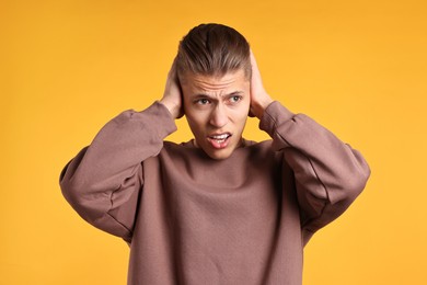 Photo of Annoyed young man covering his ears due to loud sound on orange background