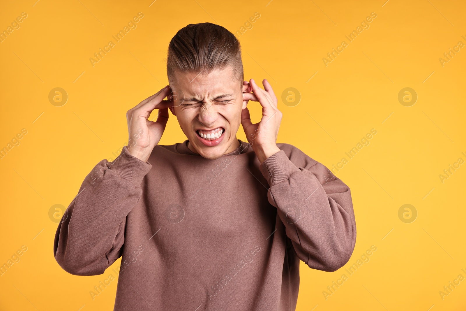 Photo of Annoyed young man covering his ears due to loud sound on orange background