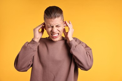 Photo of Annoyed young man covering his ears due to loud sound on orange background