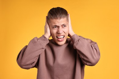 Photo of Annoyed young man covering his ears due to loud sound on orange background